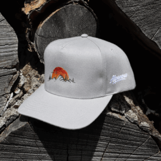 Hellgraue Alpsnow Cap mit Alpenglühen-Stick auf rustikalem Holzstapel