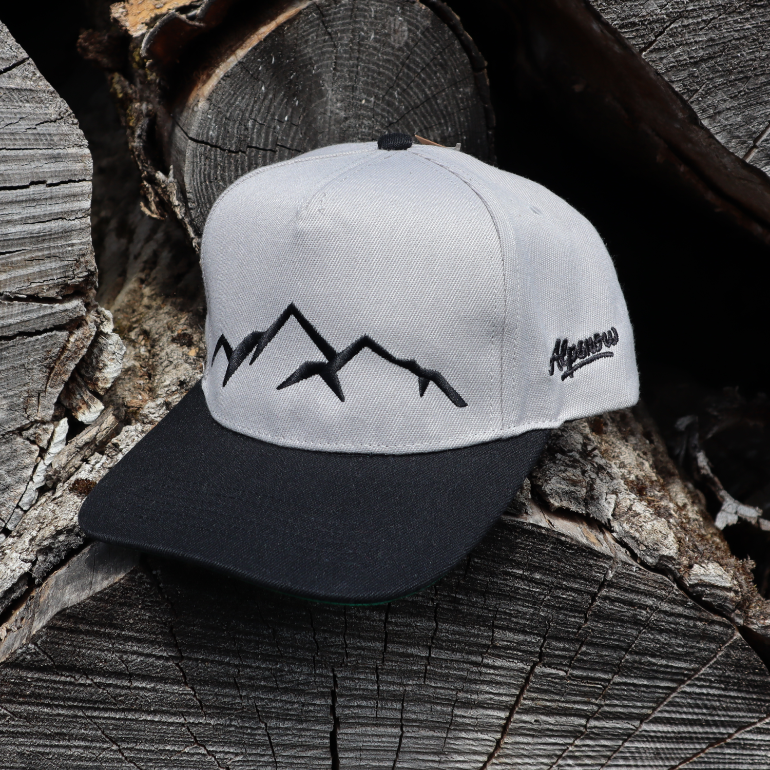 Hellgraue Alpsnow Cap mit schwarzem Schirm und schwarzem Berggipfel-Stick auf rustikalem Holzstapel