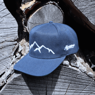 Navyblaue Alpsnow Cap mit weißem Berggipfel-Stick auf rustikalem Holzstapel