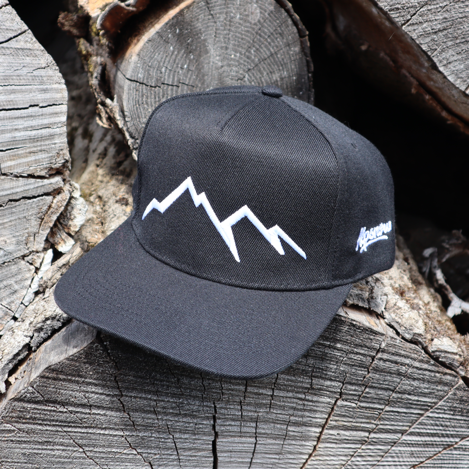 Schwarze Alpsnow Cap mit weißem Berggipfel-Stick und schwarzer Schirmunterseite auf Holzstapel