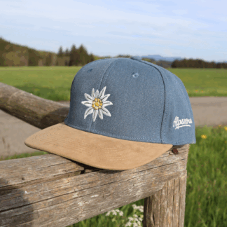 Jeansblaue Alpsnow Cap mit Edelweiß-Stick und samtigem beigem Schirm auf einem Holzzaun