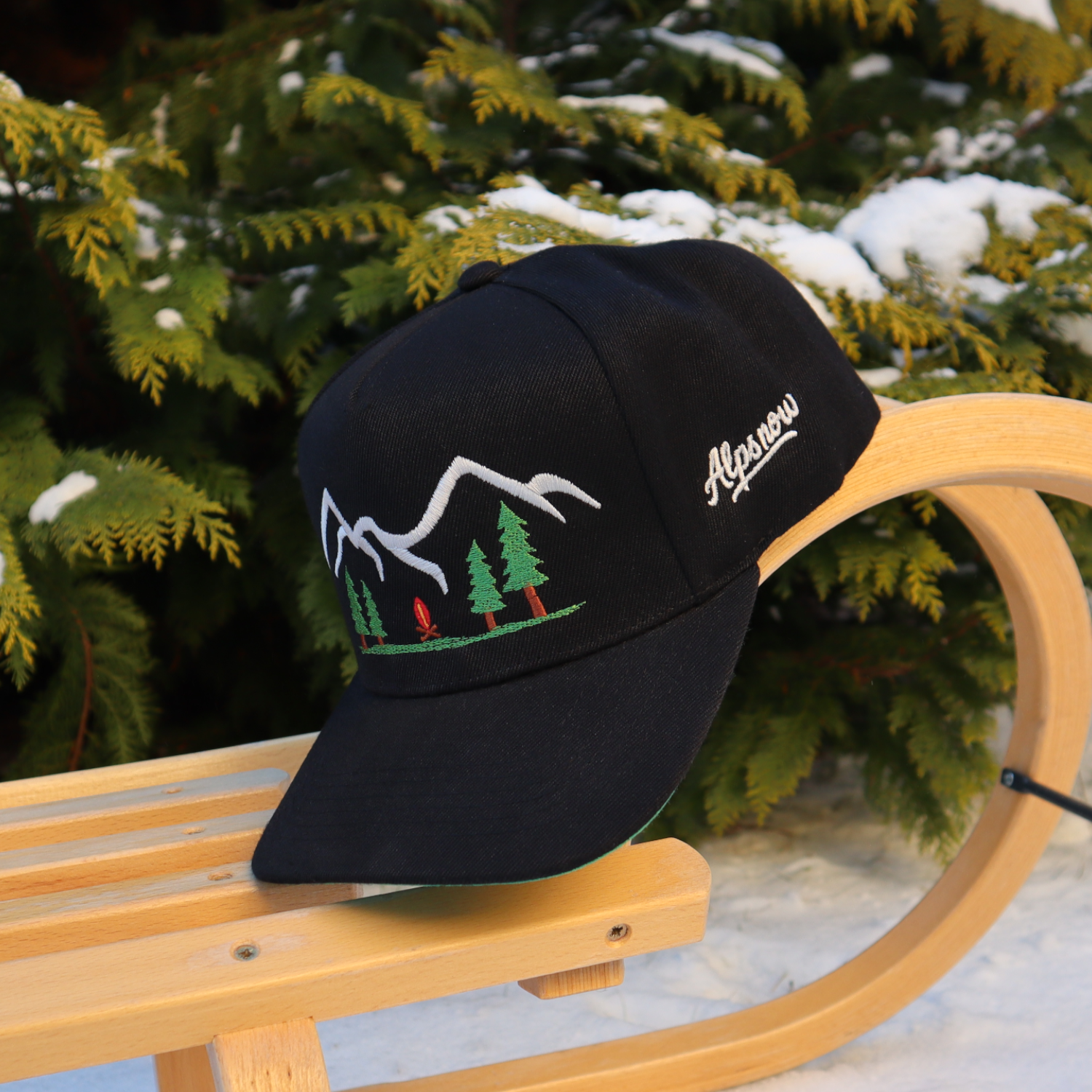 Schwarze Alpsnow Cap mit Stickerei von Bergen und Lagerfeuer auf einem Holzschlitten im Schnee