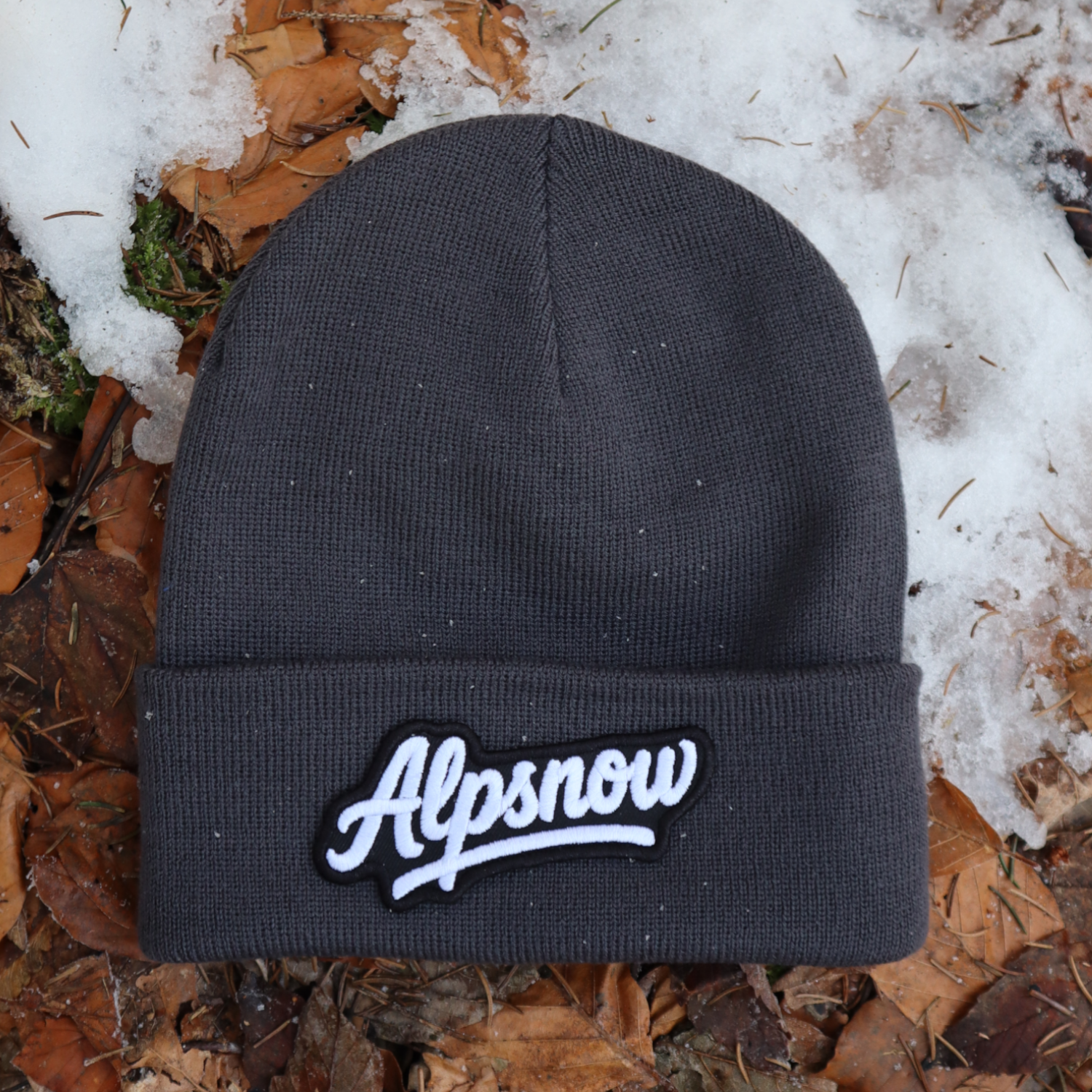 Alpsnow Mütze in modernem Grau mit einem großen, weißen Alpsnow Logo Patch auf dem Umschlag, liegend auf Waldboden und Schnee. 🏔️