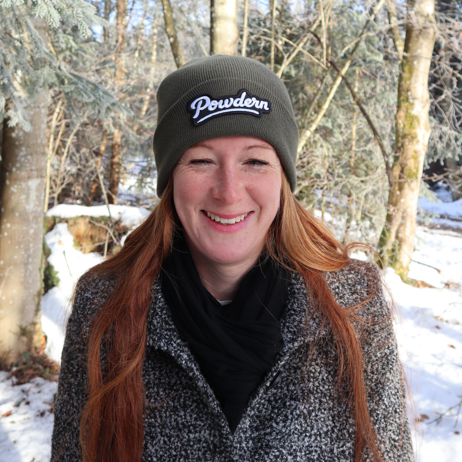 Alpsnow Mütze in modernem Grau mit einem runden, weißen Powder Patch auf dem Umschlag, getragen von einer Person in winterlicher Berglandschaft. 🏔️