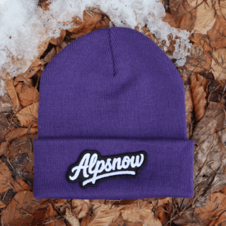 Alpsnow Mütze in intensivem Lila mit einem großen, weißen Alpsnow Logo Patch auf dem Umschlag, liegend auf Waldboden und Schnee. 🏔️
