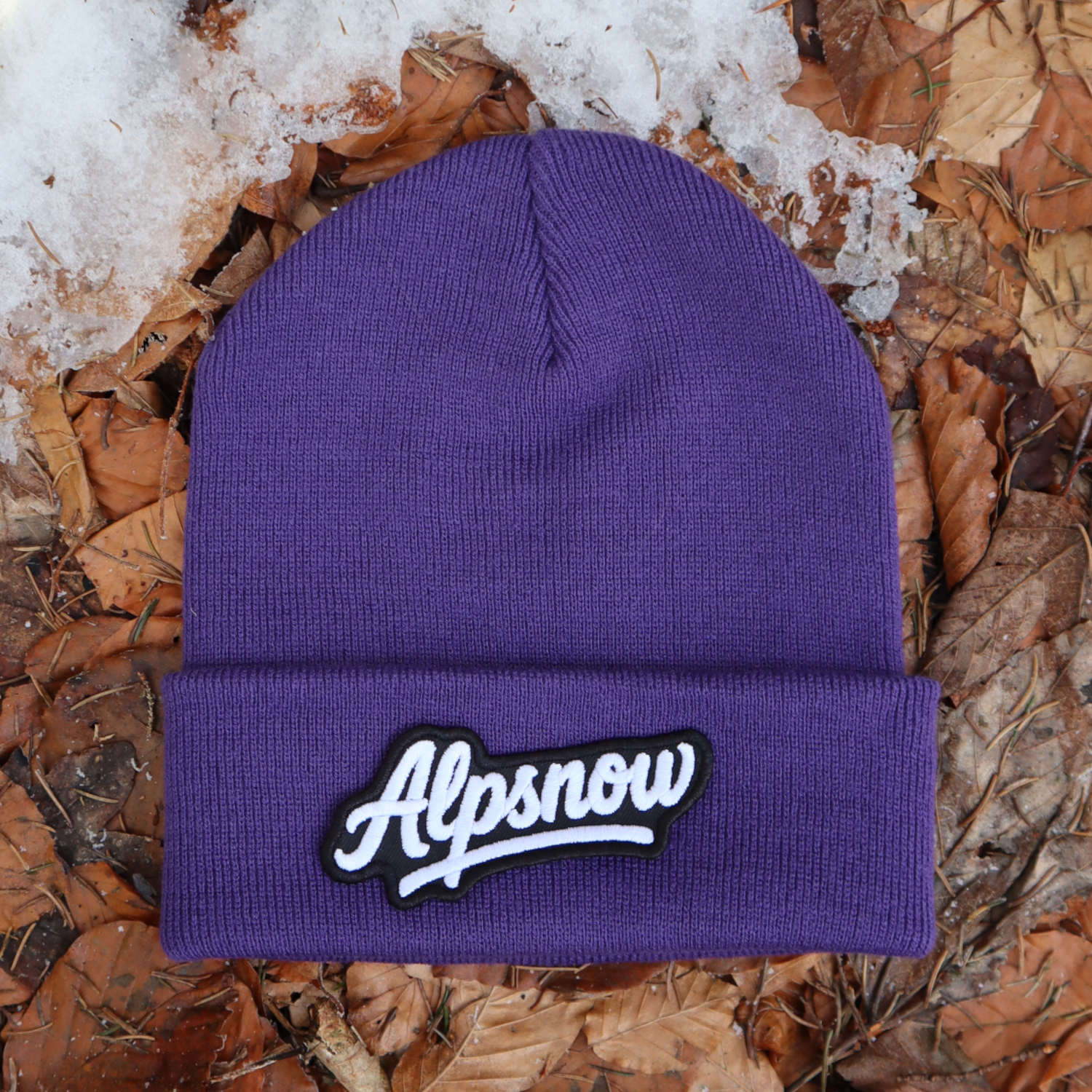 Alpsnow Mütze in intensivem Lila mit einem großen, weißen Alpsnow Logo Patch auf dem Umschlag, liegend auf Waldboden und Schnee. 🏔️