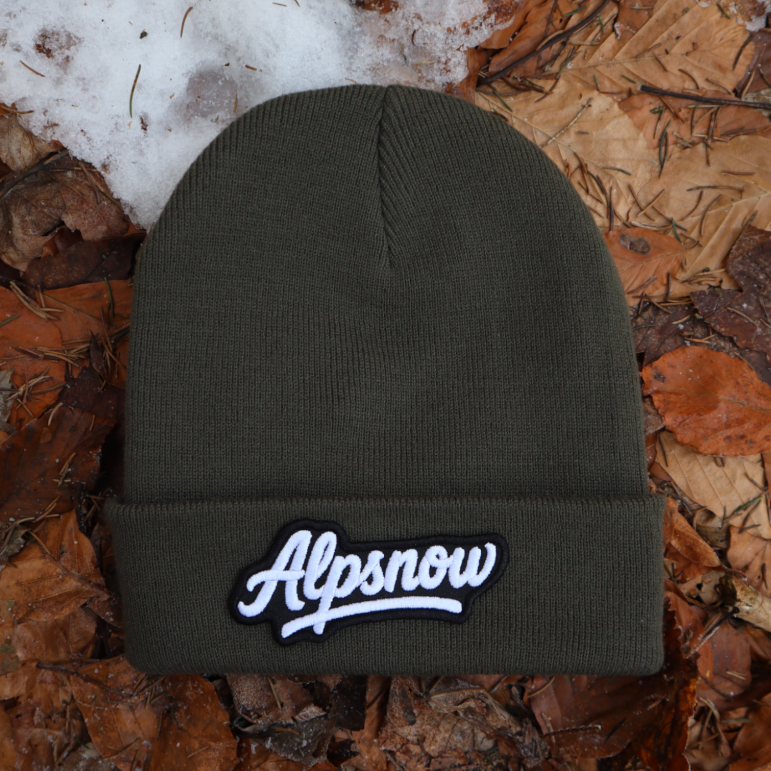 Alpsnow Mütze in modernem Oliv mit einem großen, weißen Alpsnow Logo Patch auf dem Umschlag, liegend auf Waldboden und Schnee. 🏔️