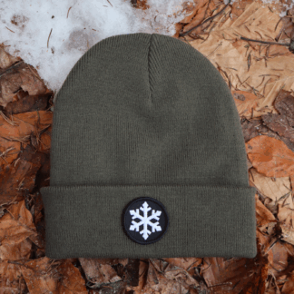 Alpsnow Mütze in modernem Oliv mit einem runden, schwarzen Patch und weißem Eiskristall auf dem Umschlag, liegend auf Waldboden und Schnee. 🏔️