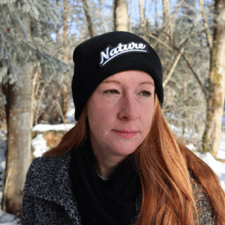 Alpsnow Mütze in tiefem Schwarz mit einem runden, beigen Nature Patch auf dem Umschlag, getragen von einer Person in winterlicher Landschaft. 🌲
