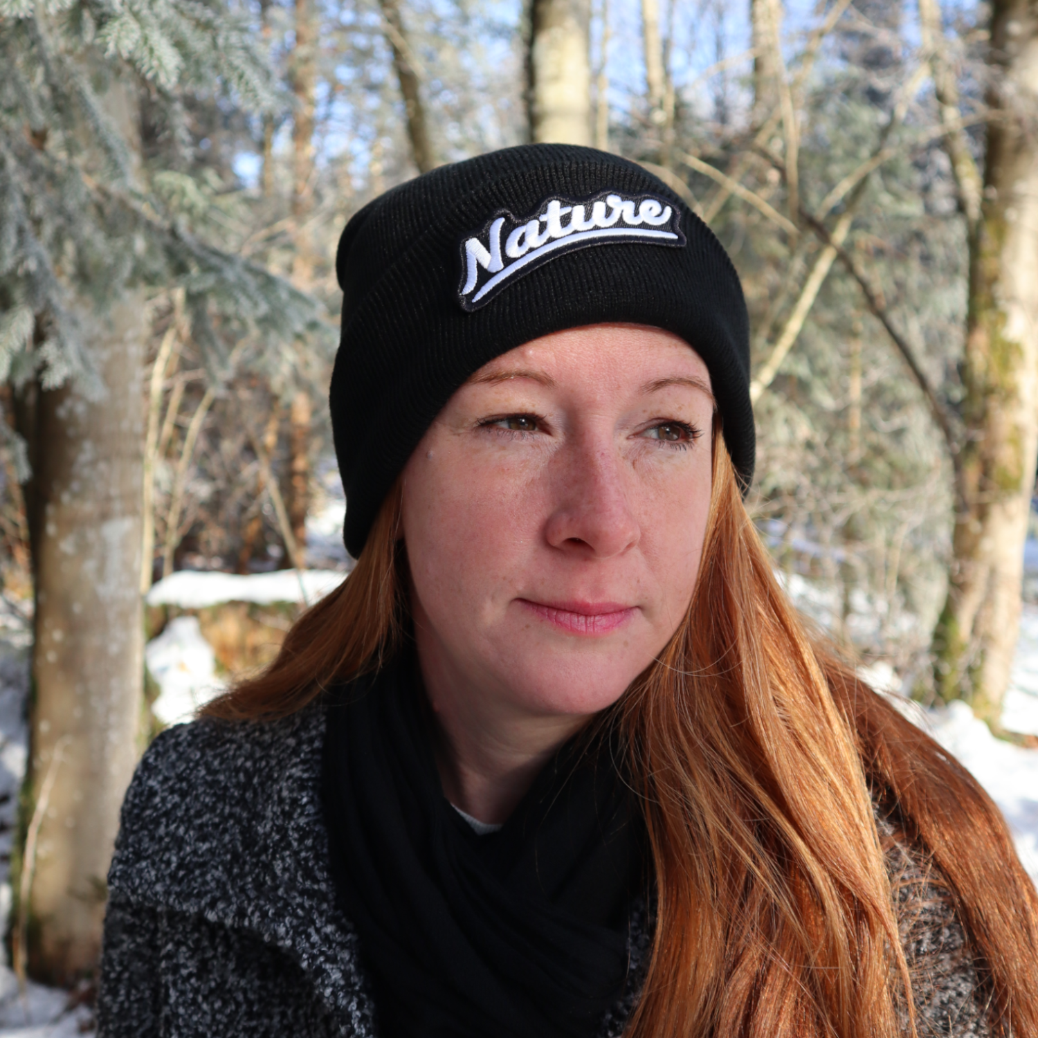 Alpsnow Mütze in tiefem Schwarz mit einem runden, beigen Nature Patch auf dem Umschlag, getragen von einer Person in winterlicher Landschaft. 🌲