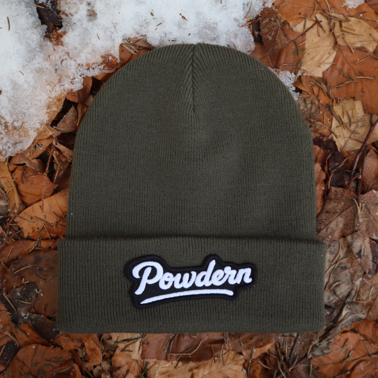 Alpsnow Mütze in modernem Oliv mit einem geschwungenen, weißen "Powdern" Patch auf dem Umschlag, liegend auf Waldboden und Schnee. 🌲