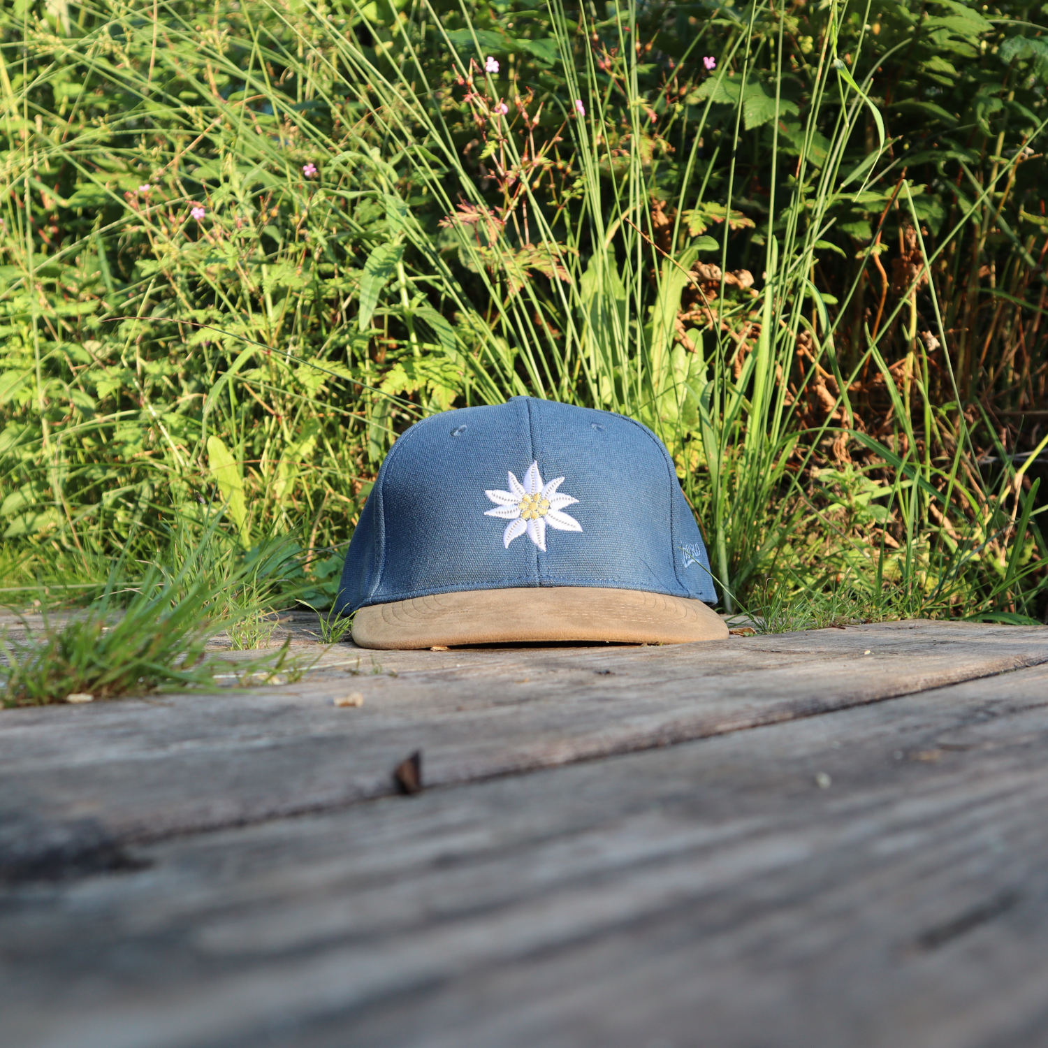 Jeansblaue Alpsnow Cap mit Edelweiß-Stick und samtigem beigem Schirm auf einem Holzzaun