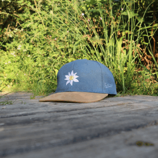 Jeansblaue Alpsnow Cap mit Edelweiß-Stick und samtigem beigem Schirm auf einem Holzzaun