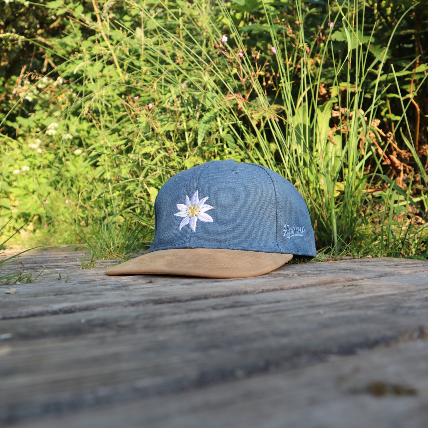 Jeansblaue Alpsnow Cap mit Edelweiß-Stick und samtigem beigem Schirm auf einem Holzzaun