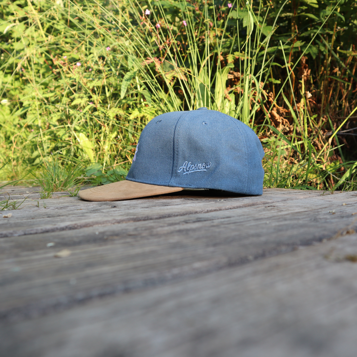 Jeansblaue Alpsnow Cap mit Edelweiß-Stick und samtigem beigem Schirm auf einem Holzzaun
