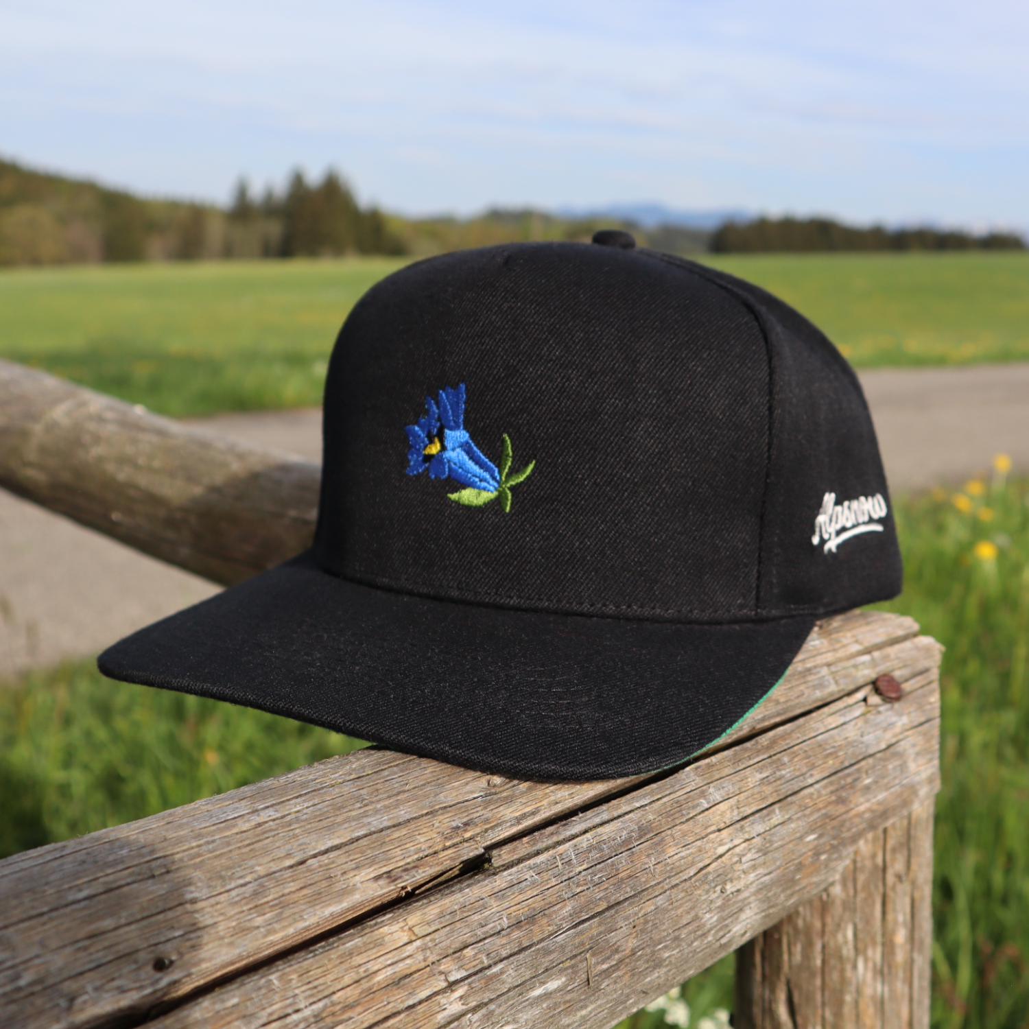 Schwarze Alpsnow Snapback Cap mit blauem Enzian-Stick auf einem Holzzaun vor einer Bergwiese