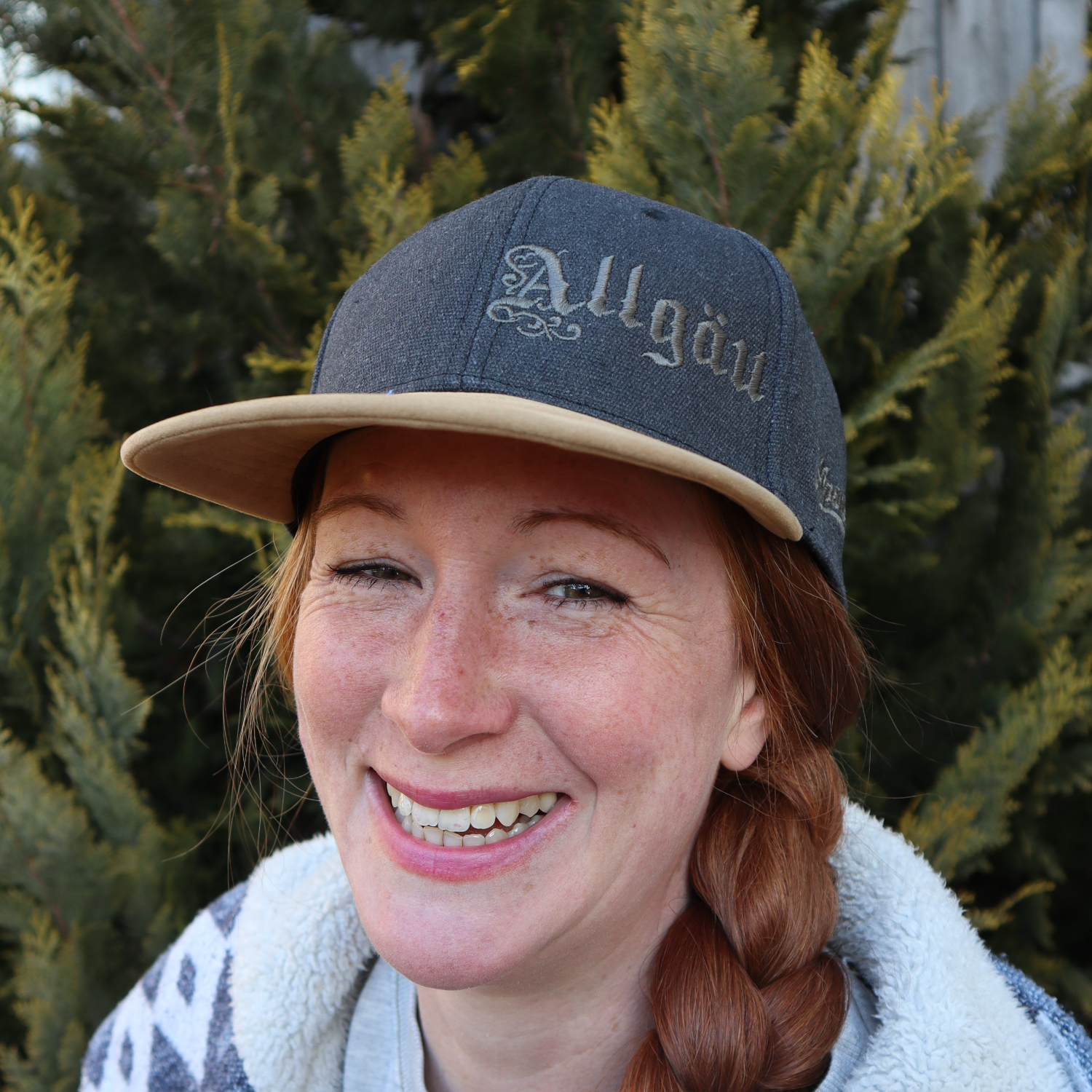 Frau mit rotem Zopf trägt die anthrazitfarbene Alpsnow Cap mit Allgäu-Stick und beigem Schirm