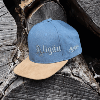 Frau mit rotem Zopf trägt die jeansblaue Alpsnow Cap mit Allgäu-Stick und beigem Schirm