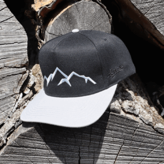 Schwarze Alpsnow Cap mit hellgrauem Schirm und weißem Berggipfel-Stick auf rustikalem Holzstapel