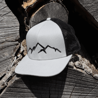 Hellgraue Alpsnow Trucker Cap mit schwarzem Netzrücken und schwarzem Berggipfel-Stick auf rustikalem Holzstapel