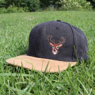 Dunkelgraue Alpsnow Cap mit Hirsch-Stick und beigem Schirm im grünen Gras
